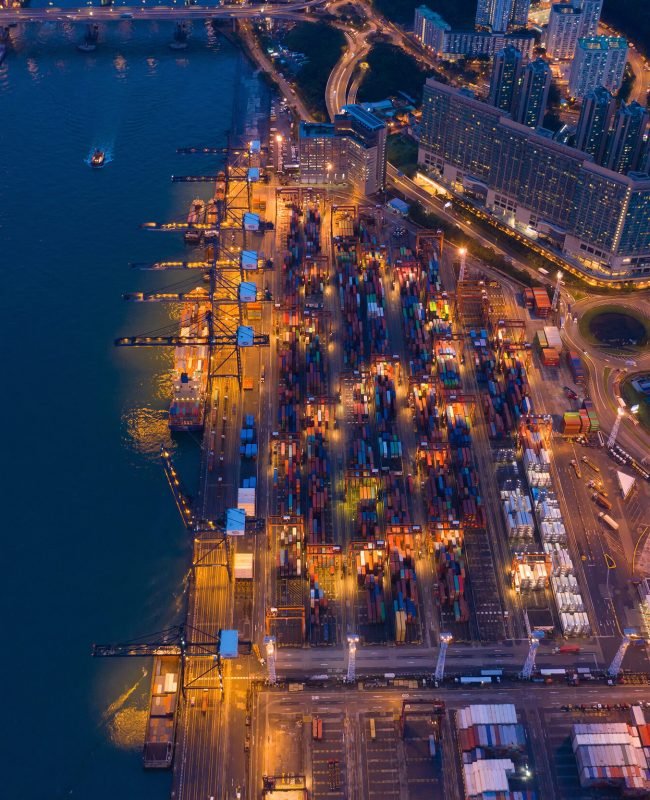 Aerial top view of container cargo ship in the export and import business and logistics international goods in urban city. Shipping to the harbor by crane in Victoria Harbour, Hong Kong City at night.